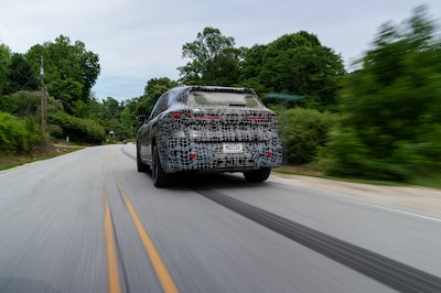 Prototipo camuflado del BMW X5 híbrido enchufable 2026 vista trasera en movimiento sobre carretera secundaria arbolada