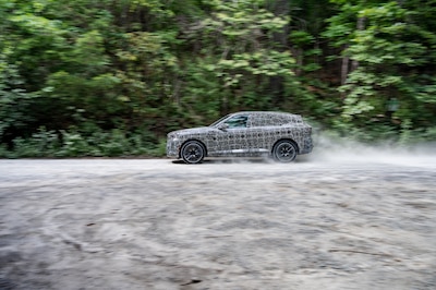 Prototipo camuflado del BMW X5 2026 vista lateral en movimiento sobre camino de tierra generando polvo durante pruebas en Spartanburg