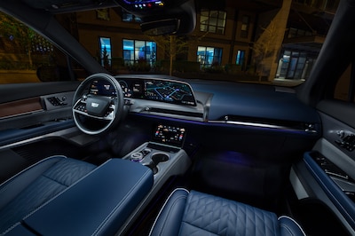 Interior del Cadillac Vistiq 2026 con iluminación ambiental azul mostrando volante, pantalla panorámica con navegación Google Maps, asientos de piel azul con costuras en patrón de diamante, consola flotante y techo panorámico de noche