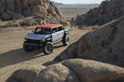 GMC HUMMER X concept pickup vista frontal tres cuartos elevada navegando paso entre formaciones rocosas en desierto árido techo rojo carrocería blanca llantas off-road de gran diámetro y salpicaderas desmontables concepto EV off-road de General Motors sin destino de producción
