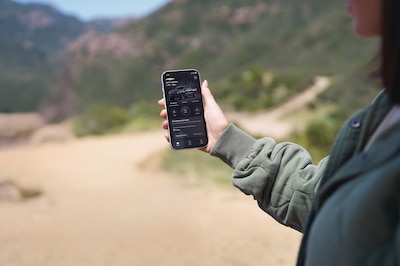 Persona sosteniendo iPhone con la aplicación MyChevrolet mostrando un Silverado 2025 con estatus del vehículo y opciones de control remoto, fondo de paisaje montañoso, app de conectividad OnStar General Motors México