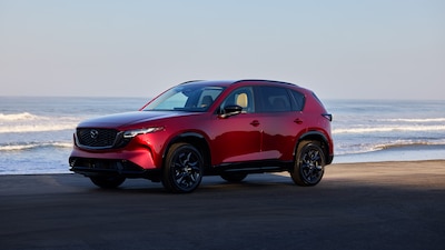 Mazda CX-5 2025 color rojo Soul Red vista tres cuartos delantera estacionado frente al mar SUV compacto de lujo tercera generación con rines negros y parrilla característica de la marca.